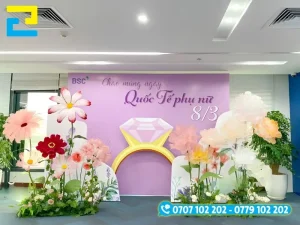 Mẫu 19: Backdrop Quốc Tế Phụ Nữ được thiết kế hình chiếc nhẫn ở giữa, có tông màu tím đẹp sang trọng, vườn hoa đẹp mắt